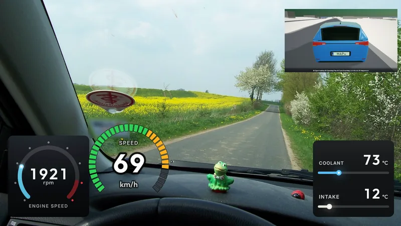 A photo through a car's windscreen onto a country road beside a rapeseed field and blossoming trees; on the dashboard engine speed (1921 rpm), a segment speedometer (69 km/h), coolant (73 °C) and intake air (12 °C), the blue 3D car on the map from behind top right.